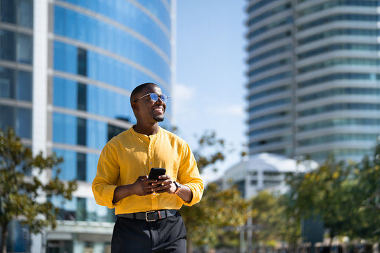 Black smiling businessman using smartphone phone, texting, standing, near glass office building. Modern African american business man works as Banker, scrolling social media, tech savvy lifestyle.