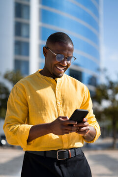 Black smiling businessman using smartphone phone, texting, closeup portrait, near glass office building. Young African american business man works as Entrepreneur, messaging colleague, modern living.