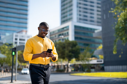 Fullbody shot, African american smiling businessman using smartphone phone, texting, near glass office building. Mid age Black business man works as Advisor, standing, browsing news feed, mobile life.