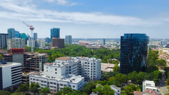 Curved blue glass skyscraper overlooks Turtle Creek corridor in Dallas. Active construction cranes rise above midrise apartment terraces beside sprawling urban forest canopies, Texas