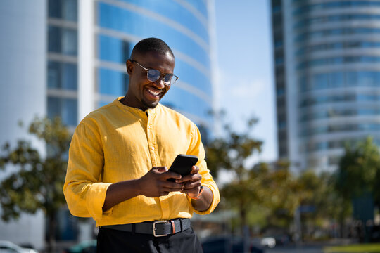 Near glass office building, Black smiling businessman using smartphone phone, texting, standing. Mature African american business man works as Sales Director, online shopping, tech savvy lifestyle.