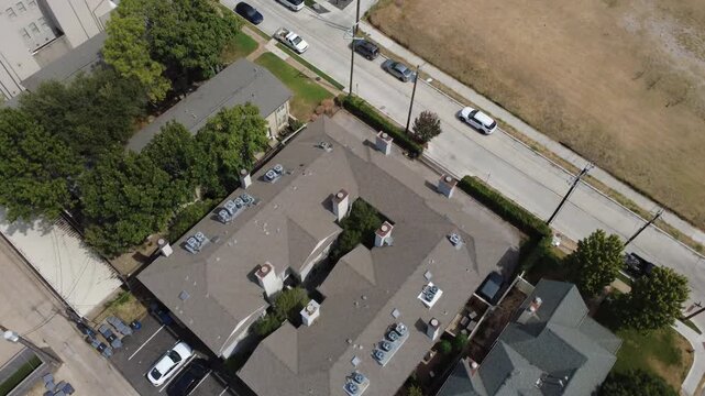 Multi tier rooflines overlook alley paving off Welborn Street in Dallas. Exposed chimney vents, rooftop air units, metal carports dot composite shingle slopes beside parked cars, Texas