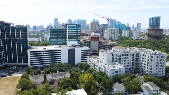 Active structural cranes rise near Welborn Street in Dallas. Midrise residential blocks with private balconies border lush urban tree canopies under hazy summer horizons, Texas