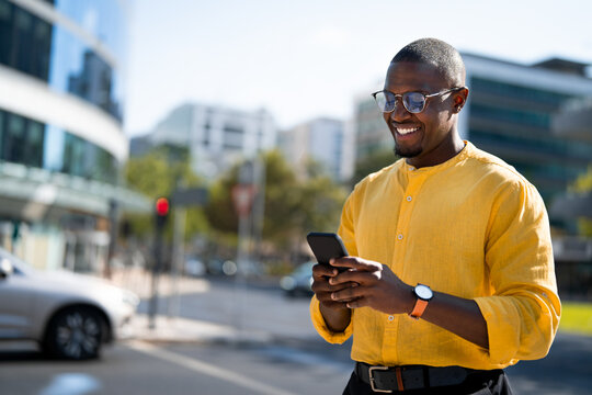 African american smiling businessman using smartphone phone, checking messages, standing at street corner, near glass office building. Young Black business man works as Manager, confident, tech savvy.