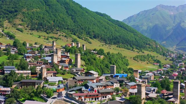 Svan defensive towers rise in forward aerial view above dense Mestia homes and mountain slopes, supporting medieval architecture, cultural tourism, family history and Georgian heritage documentaries.