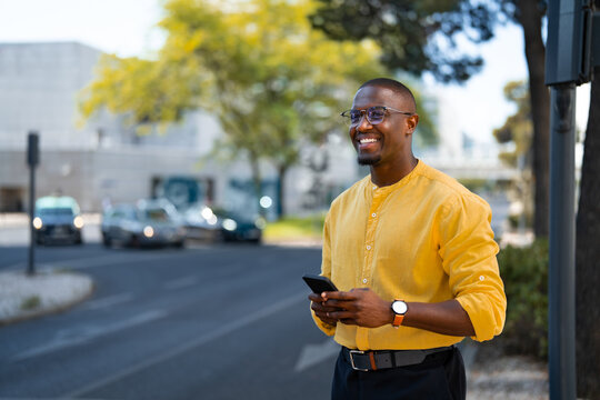 Standing casually, African american smiling businessman holding smartphone phone, looking aside, near glass office building. Mid age Black business man works as Consultant, checking calendar app.