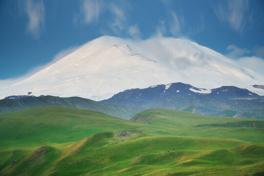 Snowy Elbrus mountain peak rises above lush green hills under a blue sky with moving clouds. High altitude volcanic landscape
