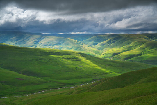 Sunlight breaks through dark storm clouds onto vast rolling green hills with a small river valley. Dramatic alpine landscape