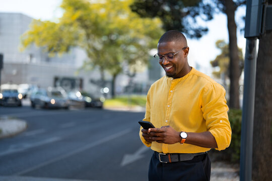 Black smiling businessman holding smartphone phone, looking aside, standing casually, near glass office building. Young African american business man works as Entrepreneur, messaging client.