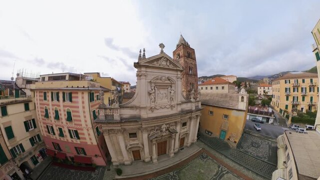 Drone orbits above historic Varazze church and patterned stone piazza framed by colorful buildings.