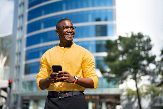 Near glass office building, African american smiling businessman using smartphone phone, texting, standing. Mid age Black business man works as Marketing Specialist, checking bank app, modern living.