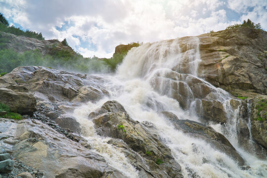 Powerful waterfall cascades down steep rocky cliffs surrounded by green trees under a cloudy sky. Alpine river stream fills