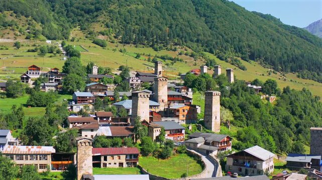 Mestia stone towers dominate forward aerial view among traditional houses beneath forested Caucasus slopes, supporting fortress architecture, heritage tourism, history education, Georgian travel media