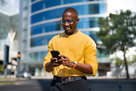 Standing, Black smiling businessman using smartphone phone, texting, near glass office building. Young African american business man works as Sales Director, video calling friend, staying connected.