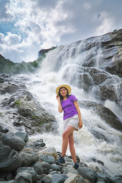Smiling woman in a straw hat stands on riverside rocks looking at camera against a big rushing waterfall. Active lifestyle portrait