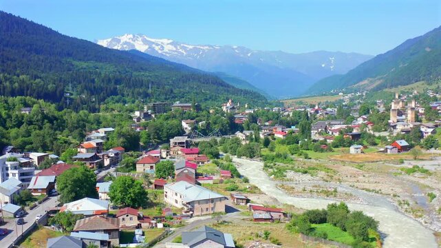 Mestia settlement fills elevated aerial view as drone retreats beside the Enguri River, mountain slopes, supporting regional tourism, urban geography, heritage travel, Georgian destination campaigns