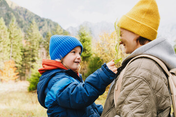 Fototapeta premium A smiling young woman and a little boy, both wearing warm beanies, hold green fern leaves in a mountain forest. Heartwarming family lifestyle concept 