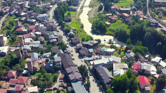 Mestia neighborhoods pass beneath forward aerial view beside a mountain river and historic tower houses, supporting city planning, river geography, heritage tourism and regional travel promotion.