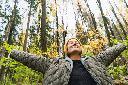 A young woman in a yellow beanie stands in a bright autumn forest with her arms stretched wide open. Joyful freedom concept expresses a deep, exhilarating connection