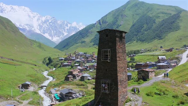 Svan tower passes in circling aerial view above Ushguli with Shkhara rising beyond the Enguri valley, supporting mountain tourism, medieval heritage, trekking promotion and Georgian travel campaigns.