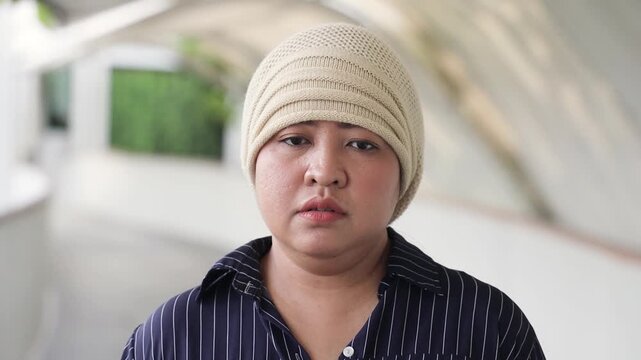 Sad and depressed middle-aged asian woman cancer patient sighing, wearing white cancer cap after suffering from serious hair loss due to chemotherapy invasive adverse event or negative side effect