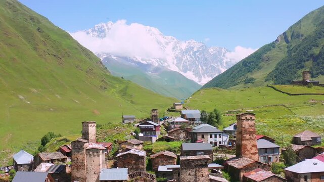 Ushguli towers recede in backward aerial view beneath Shkhara and green Caucasus slopes, supporting heritage tourism, medieval architecture, mountain geography and Georgian destination promotion.