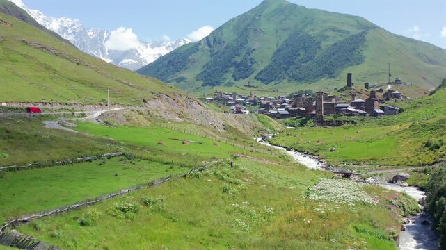 Drone advances along Enguri valley in aerial view toward Ushguli beneath green slopes and distant Shkhara, supporting trekking tourism, mountain geography, rural travel, Georgian destination campaigns