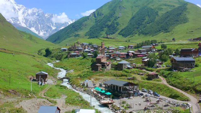 Drone advances toward Ushguli village in aerial view along the Enguri River beneath Caucasus slopes, supporting trekking routes, rural tourism, heritage travel, Georgian mountain destination campaigns