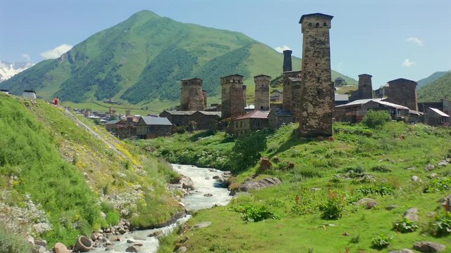 Drone advances along Enguri River in aerial view beside Chazhashi towers and historic stone houses, supporting river geography, medieval heritage, rural tourism and Upper Svaneti destination promotion