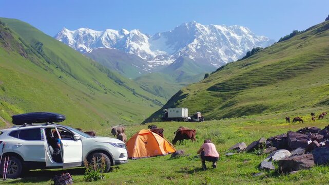 Campers rest in hovering aerial view beside an SUV and orange tent in the Shkhara valley near Ushguli, supporting self-drive tourism, outdoor recreation, trekking access and Georgian travel campaigns.