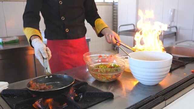 Unrecognizable chef holding two pans and cooking tasty flambe style dish in modern kitchen of hotel. Male cook working with different ingredients in cuisine. View on cooking process. Slow motion