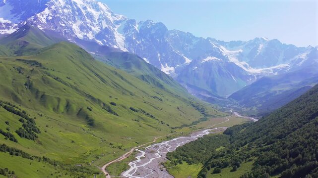 Drone flies forward above Enguri valley toward Shkhara massif near Ushguli, revealing braided river channels and alpine slopes for trekking tourism, glacier geography, Georgian destination campaigns
