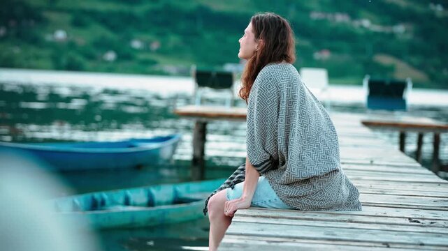 Young woman wrapped in a warm blanket sitting on a wooden pier by a scenic mountain lake, enjoying the peaceful atmosphere and breathtaking mountain views.