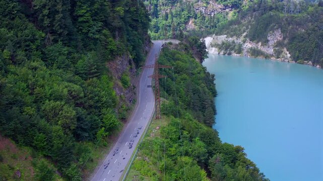 Drone hovers above Enguri Reservoir beside a narrow mountain road and forested shoreline in Svaneti, Georgia, supporting road tourism, reservoir geography, transport planning and destination media.
