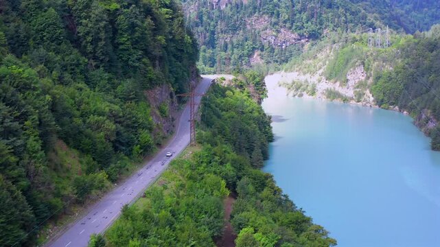Drone flies forward along Enguri Reservoir road beside turquoise water and steep wooded slopes in Svaneti, Georgia, supporting scenic driving, route planning, mountain tourism and transport media.