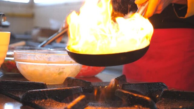 Unrecognizable chef holding two pans and cooking flambe style dish in kitchen of hotel. Male cook tossing and frying vegetables in flaming skillet on stove. Concept of cooking. Slow motion Side view
