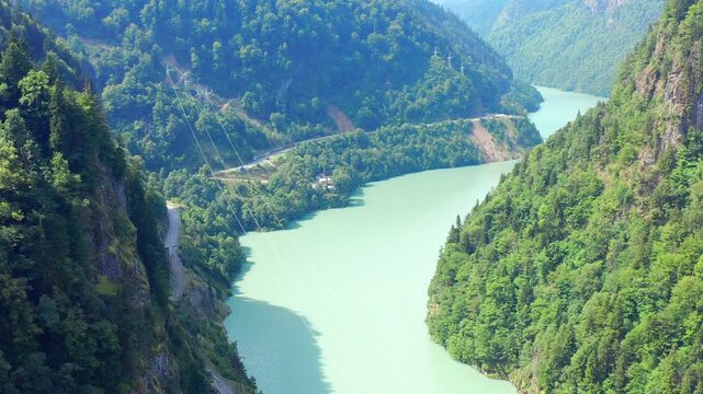 Drone flies forward above Enguri Reservoir through forested mountain terrain with shoreline roads and power lines, supporting hydropower geography, infrastructure planning and Georgian travel media.