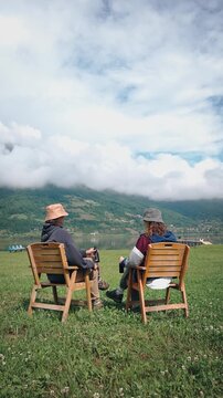 Two female travelers sitting on wooden chairs by a scenic mountain lake, drinking beer and enjoying breathtaking mountain views during a relaxing outdoor vacation.