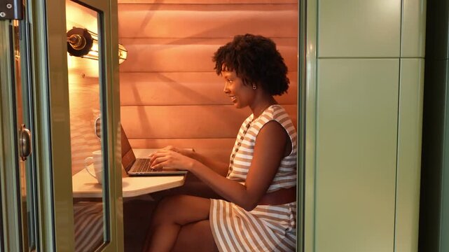Young african american businesswoman working on a laptop computer inside a private pod in a modern coworking office space, then smiling
