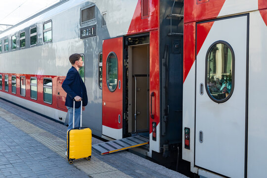 Man carrying a yellow suitcase walks up the ramp to board a double-decker train at a modern railway station, with the train's exterior featuring red and gray colors