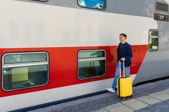 Young man with yellow suitcase walks beside a red and gray train at a railway station, preparing for travel on a sunny day. Business trip and vacation concept.