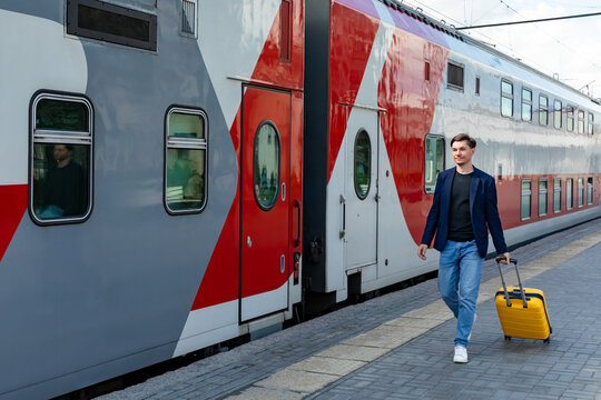 Young man in a blue blazer and jeans pulls a yellow suitcase while walking next to a modern train at a railway station, highlighting urban travel and the dynamic atmosphere of transportation