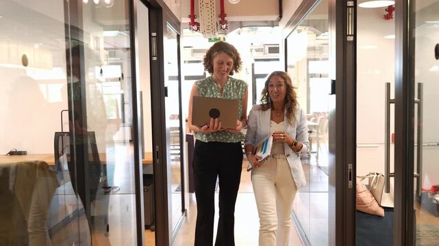 Two mature businesswomen having a conversation while walking down a modern office corridor. One colleague holds a laptop while the other holds documents