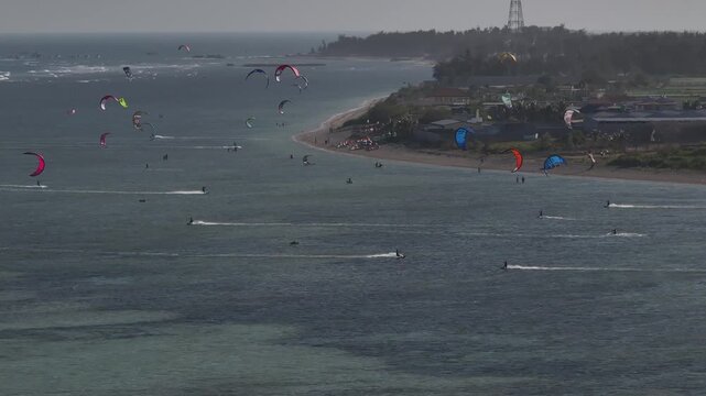 Aerial drone view of kite surfers riding the waves at My Hoa Beach in Vinh Hai, Khanh Hoa, Vietnam, showcasing a spectacular coastal landscape under bright sunlight.

