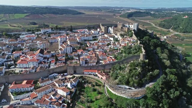 Aerial view of the historic walled town of Obidos with its medieval castle, red-tiled roofs, and surrounding green landscape in Obidos, Leiria District, Portugal.
