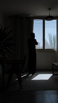 Silhouette of a distressed man standing alone by a window and covering his face, overwhelmed by emotional stress, worry and a difficult realization in a dark dramatic room, vertical