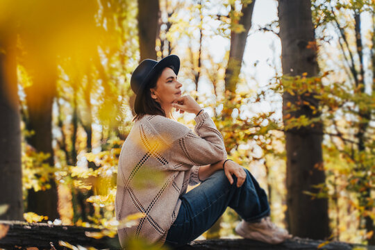 Smiling mid-adult perimenopausal Caucasian woman in a felt hat sitting on a log in the autumn forest.