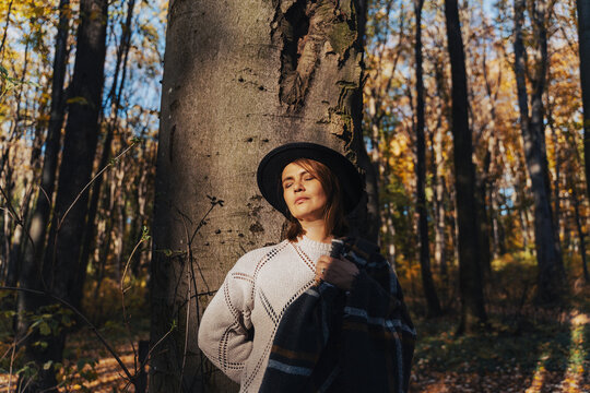 Peaceful mid-adult woman in a felt hat standing by a tree trunk in autumn forest with closed eyes.