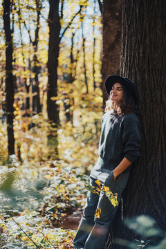Vertical portrait of a dreamy young woman in a hat leaning against a tree trunk in autumn forest.