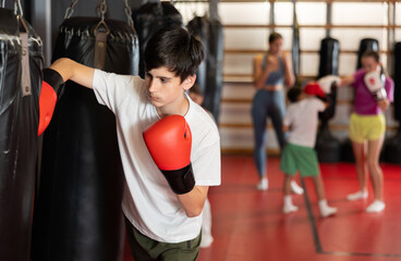 Fototapeta premium Boy in red boxing gloves stands near a punching bag. Child in boxing training learns punches for self-defense
